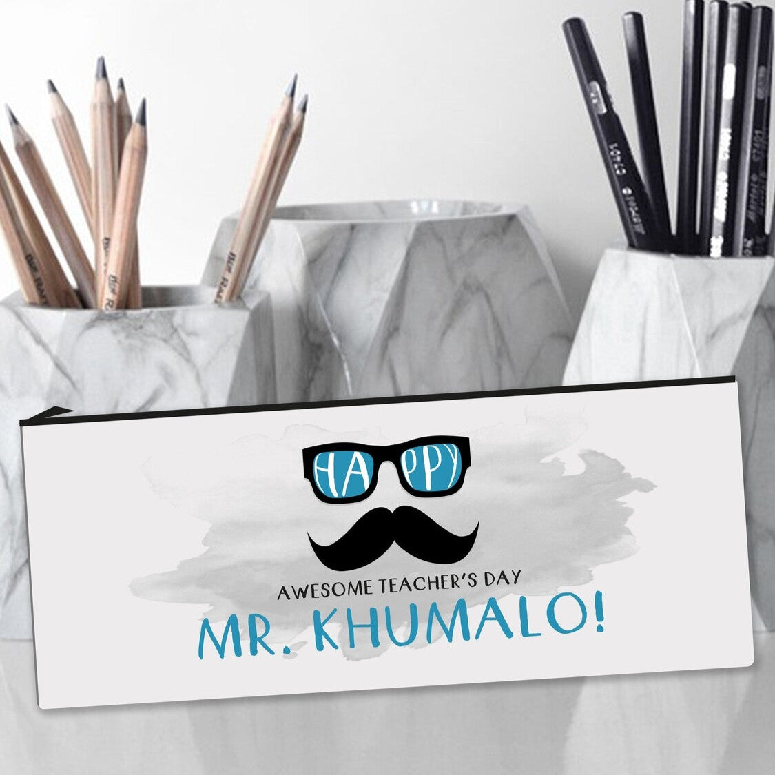 Personalized pencil case that says "MR. KHUMALO!" with "AWESOME TEACHER'S DAY!" and a graphic of sunglasses and a moustache, placed near marble pencil holders containing wooden and black pencils.