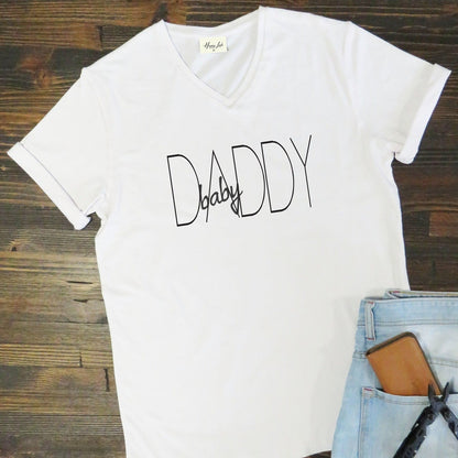 Men’s T-Shirt “Baby Daddy” from Happy Look | South Africa – A white V-neck T-shirt with black minimalist text reading “baby DADDY.” Displayed flat on a wooden background with a pair of light blue jeans, phone, and keys beside it.