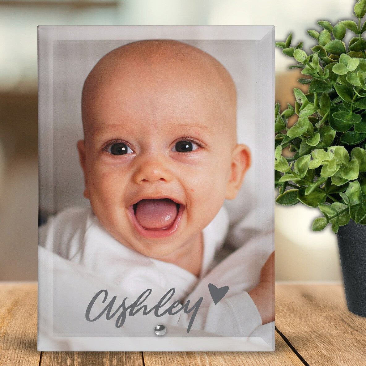 Personalised Glass Photo Tile | Happy Look South Africa - Personalised glass photo tile featuring a smiling baby with the name “Ashley” and a small heart printed on the front. The tile stands on a wooden surface beside a green plant, softly lit in a cozy home setting.