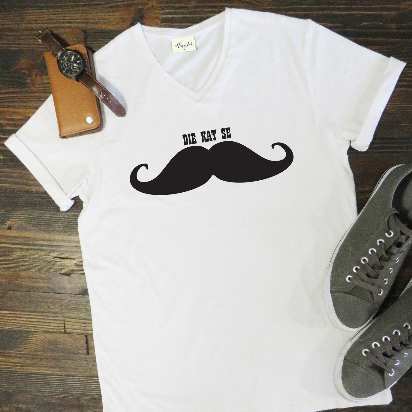 White t-shirt with a black mustache graphic and text on a wooden surface with shoes and a watch.