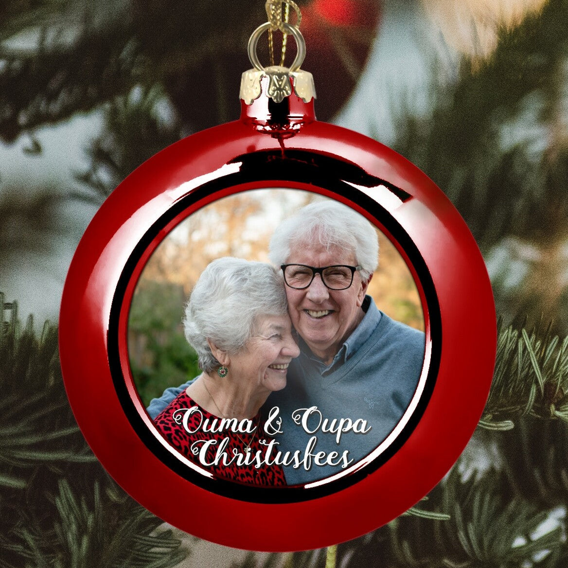 Custom Grandparent Photo Bauble "Ouma & Oupa Christussfees" from Happy Look | South Africa – A glossy red Christmas ornament hanging from a tree. The center displays a photo of a loving elderly couple, overlaid with the text "Ouma & Oupa Christussfees" (Grandma & Grandpa Christmas). A heartwarming family gift.