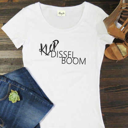 White t-shirt with 'Klop Dissel Boom' text on a wooden surface with jeans and sandals.