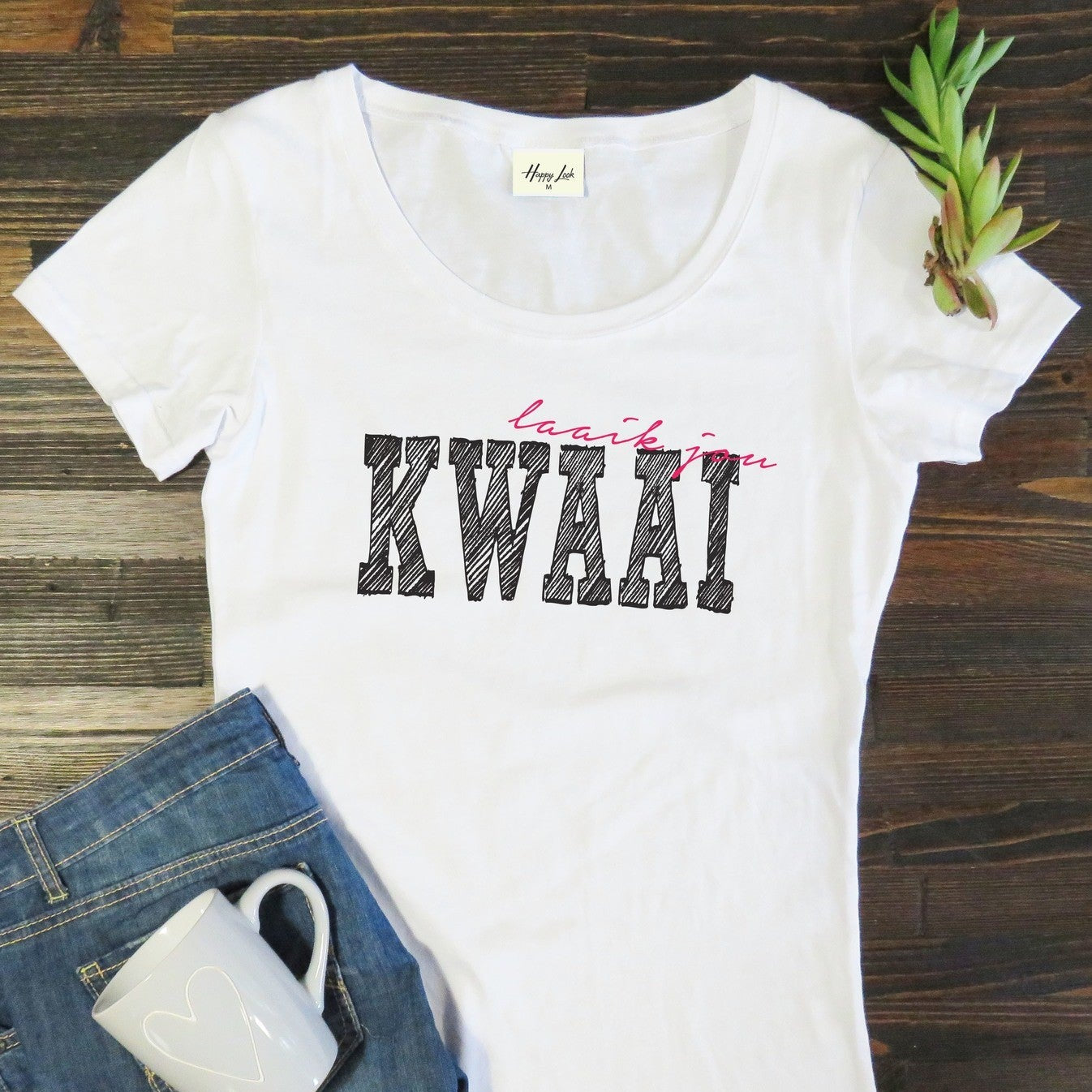 White t-shirt with 'Laai jou KWAAI' text on a wooden surface with jeans and a mug.