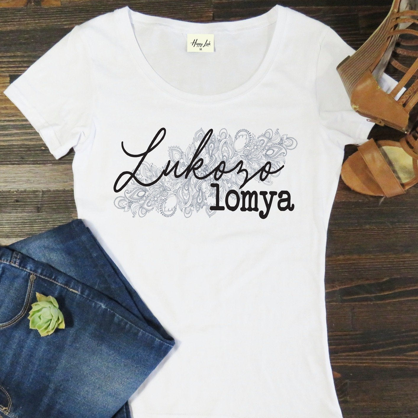White t-shirt with 'Lukozo lomya' text on a wooden surface with jeans and sandals.