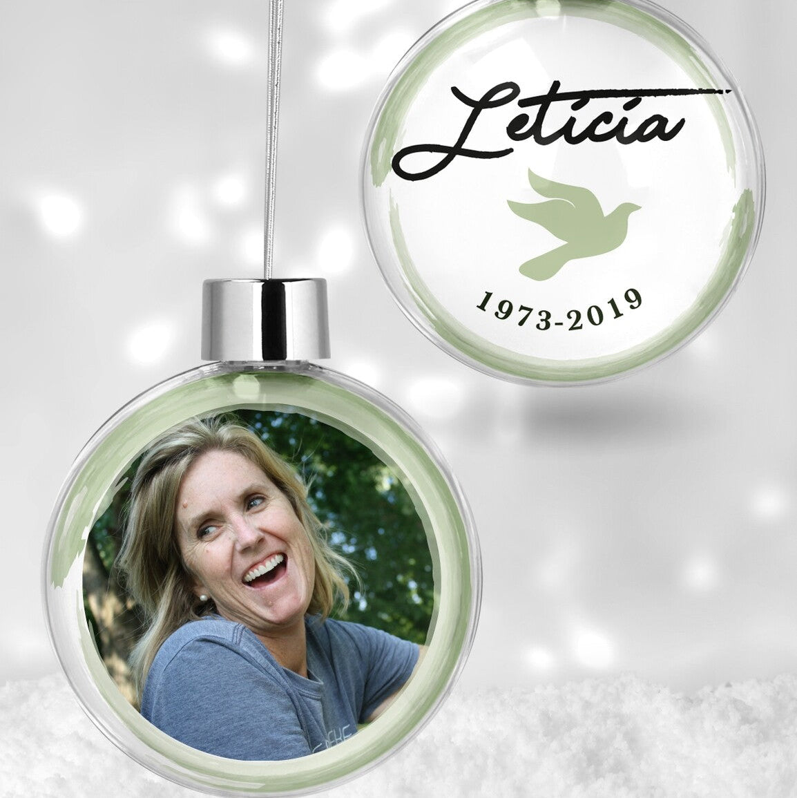 Custom Memorial Christmas Bauble "Letícia 1973-2019" from Happy Look | South Africa – A clear hanging Christmas bauble set. One bauble has a photo of a smiling woman, and the other displays the name "Letícia," a dove, and "1973-2019" against a soft green background.