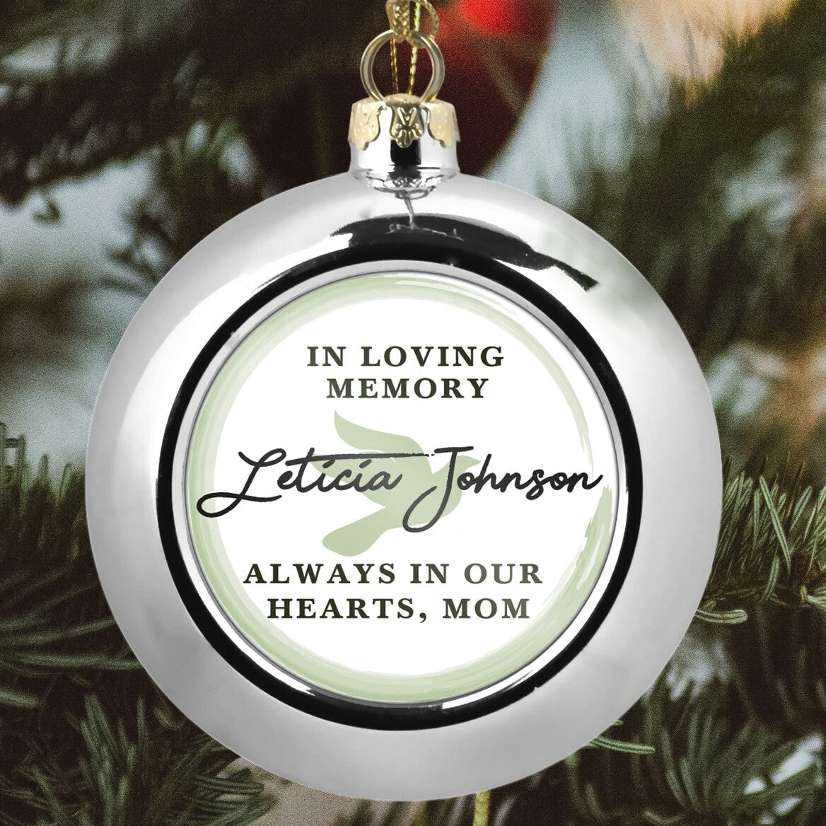 Custom Memorial Christmas Bauble "In Loving Memory Leticia Johnson" from Happy Look | South Africa – A glossy silver Christmas ornament hanging from a tree. The center features a memorial text circle: "IN LOVING MEMORY," the name "Leticia Johnson" in script, a dove, and "ALWAYS IN OUR HEARTS, MOM."