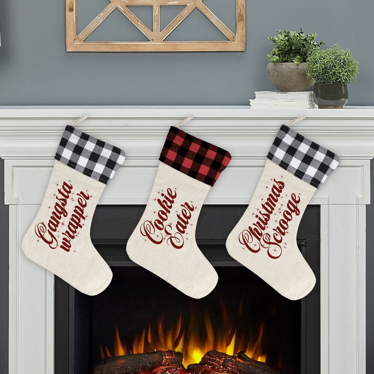 Humorous Christmas Stockings "Gangsta Wrapper, Cookie Eater, Christmas Scrooge" from Happy Look | South Africa – Three hanging stockings on a mantel with cream bodies and buffalo plaid cuffs (alternating black and red). Each is printed with a funny red script phrase: "Gangsta Wrapper," "Cookie Eater," and "Christmas Scrooge."