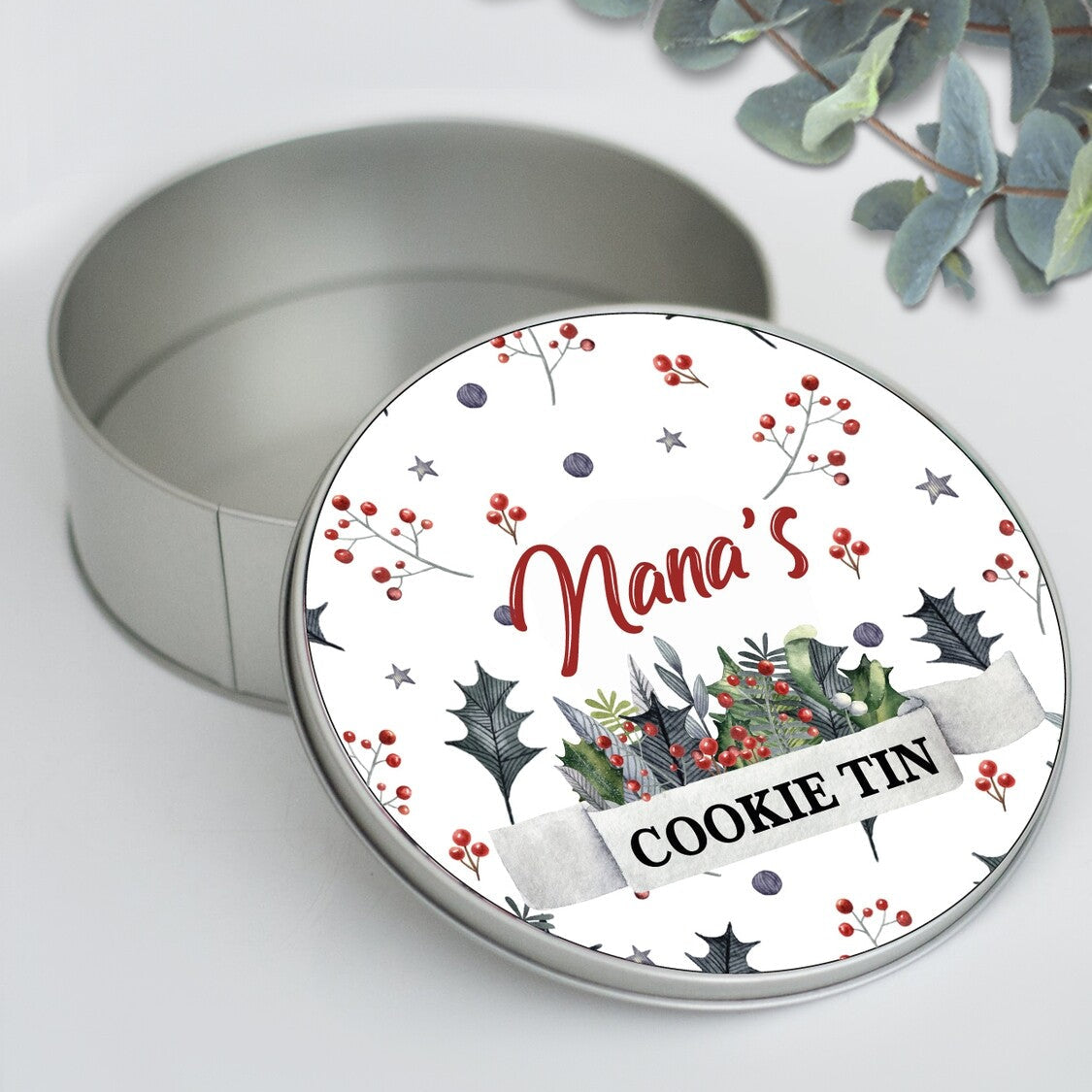 Round Personalized Christmas Cookie Tin "Nana's COOKIE TIN" from Happy Look | South Africa – An open round silver tin with its lid leaning against it. The lid features a watercolor design of holly and berries around the script text "Nana's" and a banner with "COOKIE TIN."