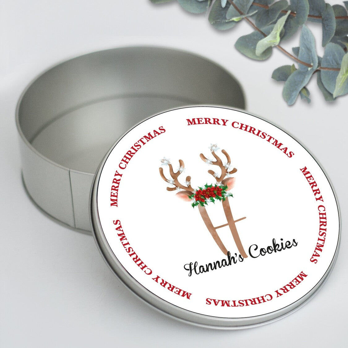 Round Personalized Christmas Cookie Tin "Hannah's Cookies" with Reindeer Antlers from Happy Look | South Africa – An open round silver tin with its lid leaning against it. The lid features a simple wreath of "MERRY CHRISTMAS" and a design of reindeer antlers decorated with holly, with the script text "Hannah's Cookies."