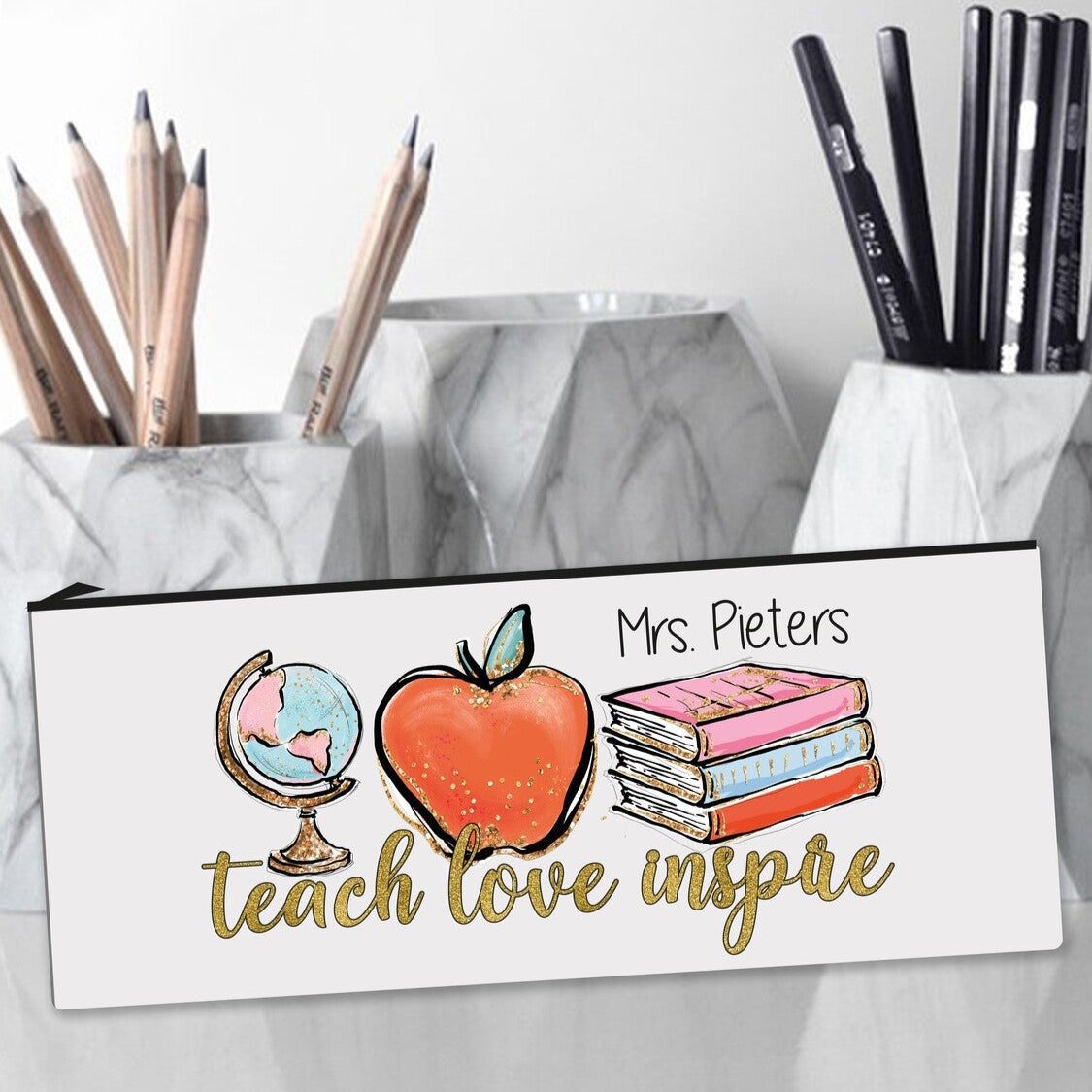 Personalized pencil case with the name "Mrs. Pieters," displaying the phrase "teach love inspire" alongside illustrations of a globe, an apple, and stacked books, next to marble pencil holders filled with wooden and black pencils