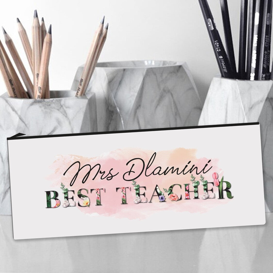 Personalized pencil case featuring the name "Mrs. Dlamini" with "BEST TEACHER" in decorative floral letters, placed next to marble pencil holders filled with wooden and black pencils.